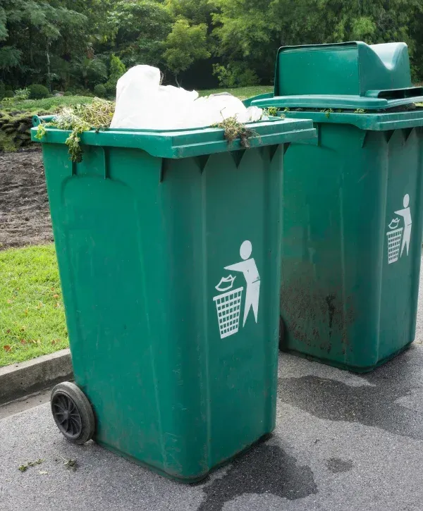 Two green garbage dumpsters standing on a paved surface, the front one overflowing with trash.