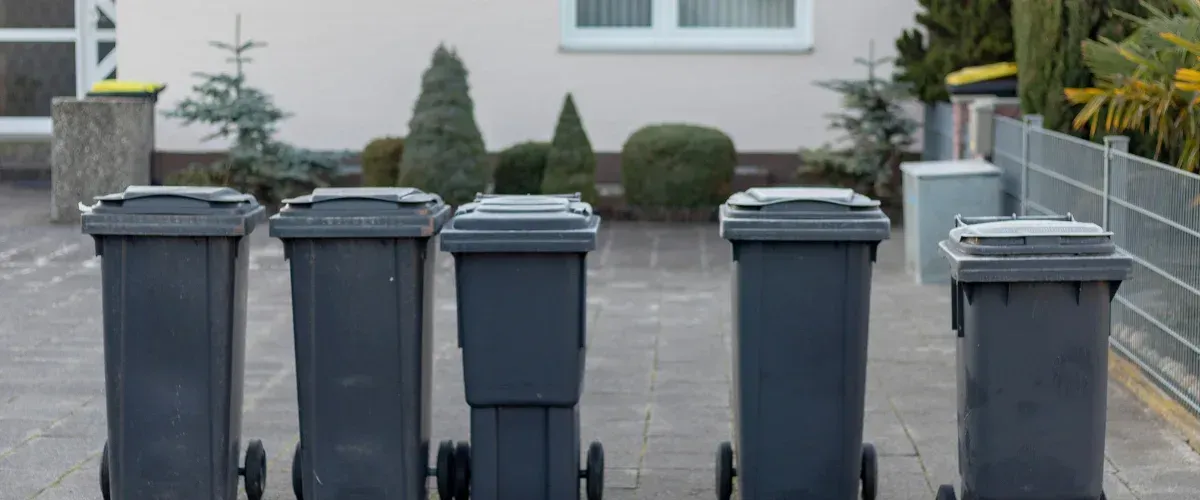 A row of five dark gray trash cans with lids, lined up on a paved surface in front of a house.