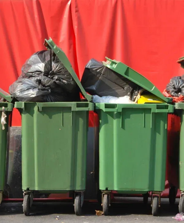 Several green trash bins overflowing with black garbage bags and trash against a bright red backdrop.