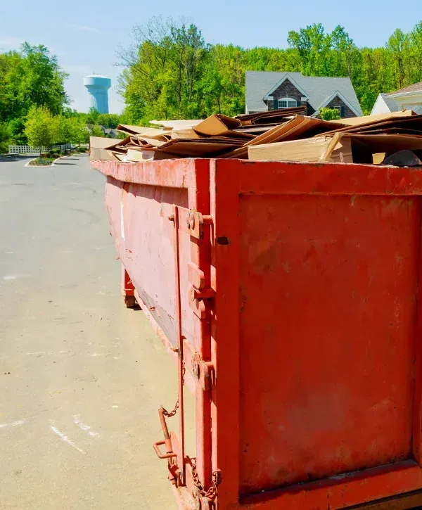 A long orange dumpster overflowing with cardboard waste, parked on asphalt with houses and trees in the background.