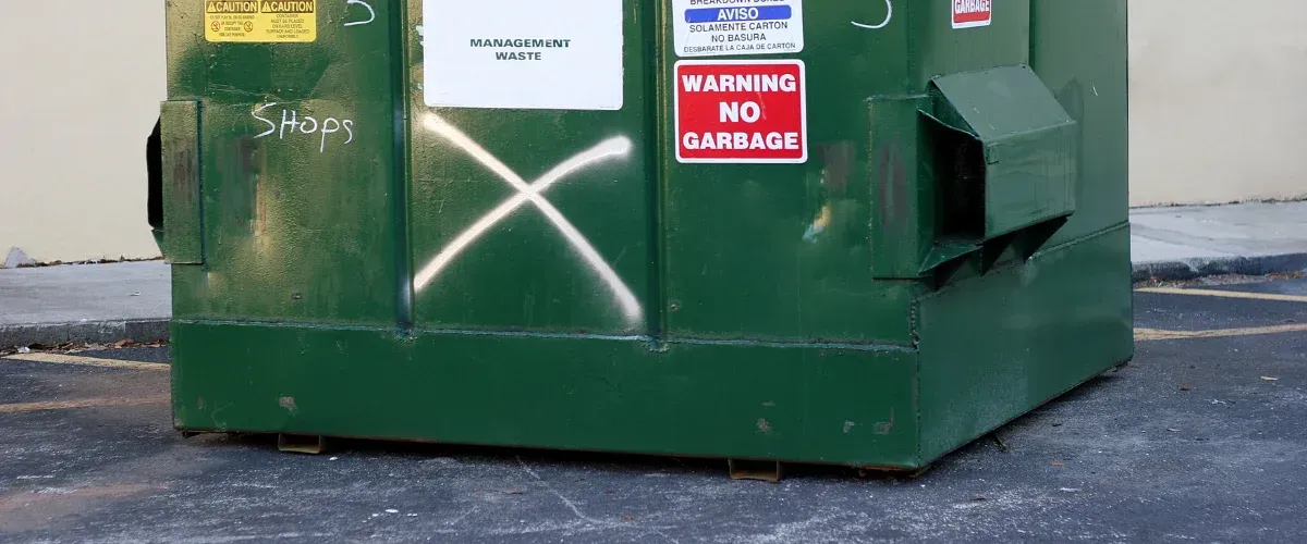 A large green dumpster with a white painted 'X' and a "Warning No Garbage" sign on its side.