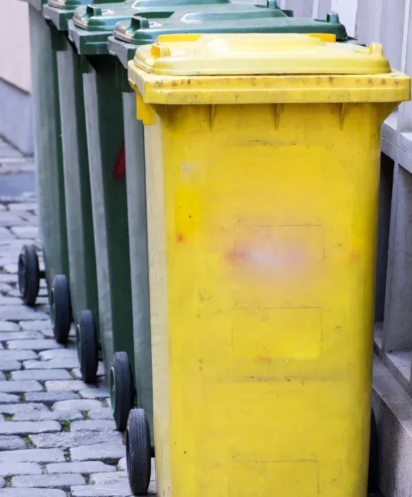 A row of green and yellow recycling bins with wheels lined up on a cobblestone street next to a building.