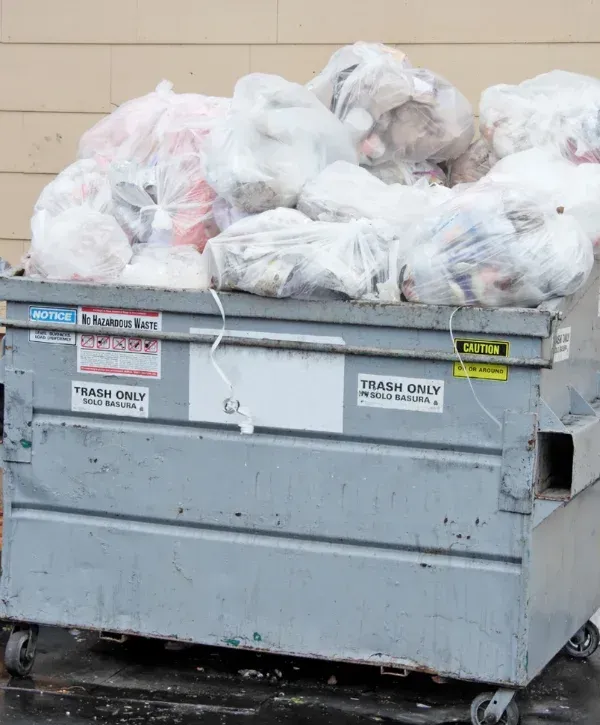 A large gray dumpster overflowing with numerous white plastic bags filled with trash, with a beige building in the background.