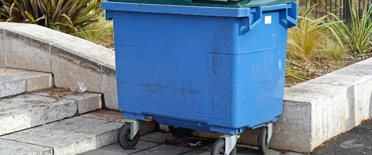A large green dumpster with a white painted 'X' and a "Warning No Garbage" sign on its side.