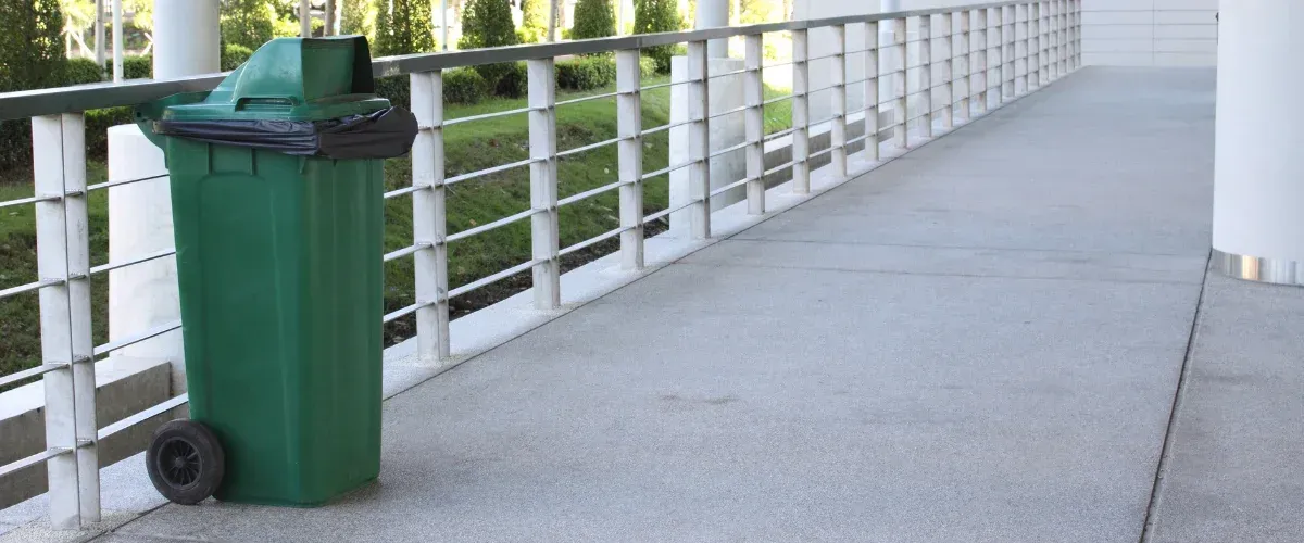 A green wheeled trash bin with a black liner on a concrete walkway next to a modern metal railing.