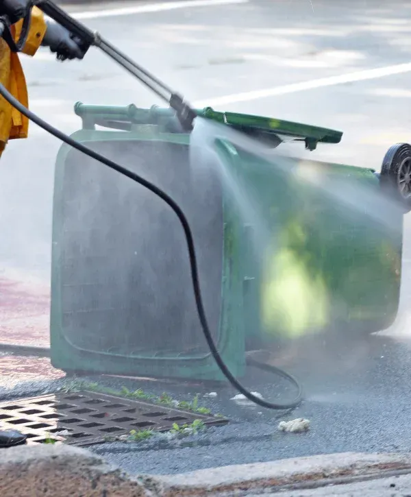 A person in yellow protective gear uses a pressure washer to clean a green wheelie bin lying on its side on asphalt.