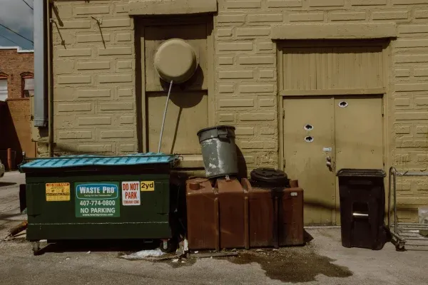 A back alley with a green dumpster, brown containers, and a trash can against a textured yellow brick wall.