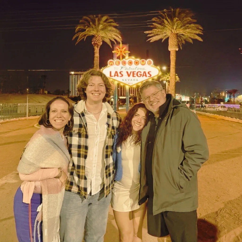 A candid, softly lit 3:2 family portrait of John, Maria, Catarina, Juliano, and their golden retriever Ava, all sitting together on a beige linen sofa in a sunlit living room with sand-toned walls and minimal decor. Everyone is smiling warmly, dressed in cozy, neutral clothing, radiating togetherness and joy.