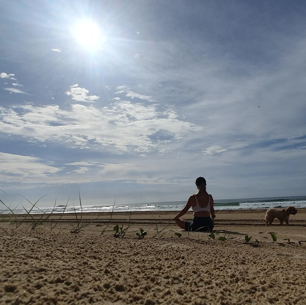 Woman sitting on a beach, crossed legged meditating while a facing the sun while a cavodle wanders by. 