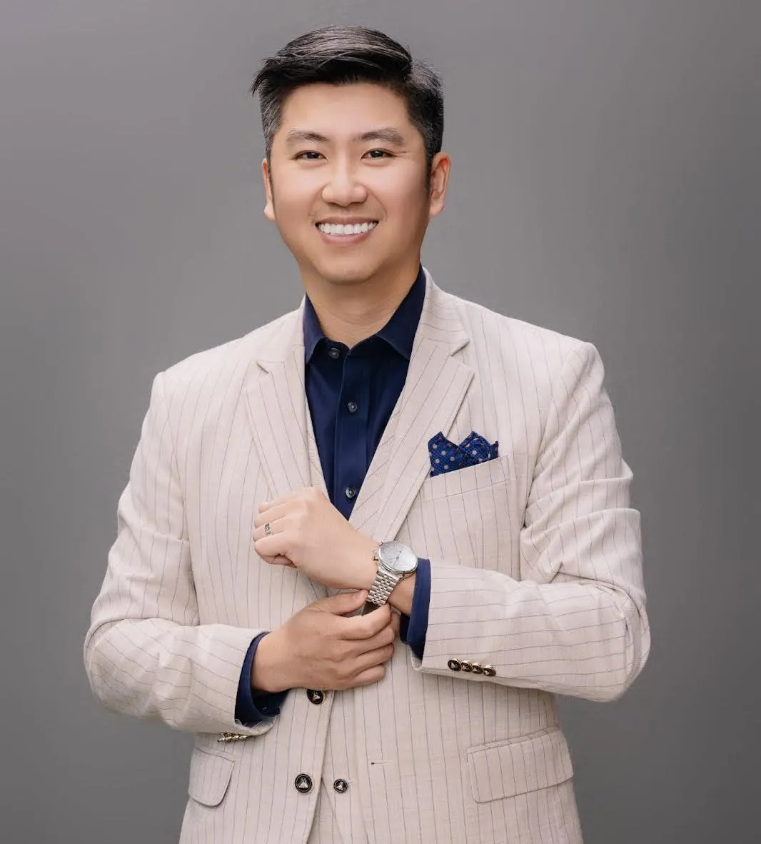 A man in a stylish cream pinstripe suit and navy shirt, with a blue pocket square, adjusts his watch, against a gray background.
