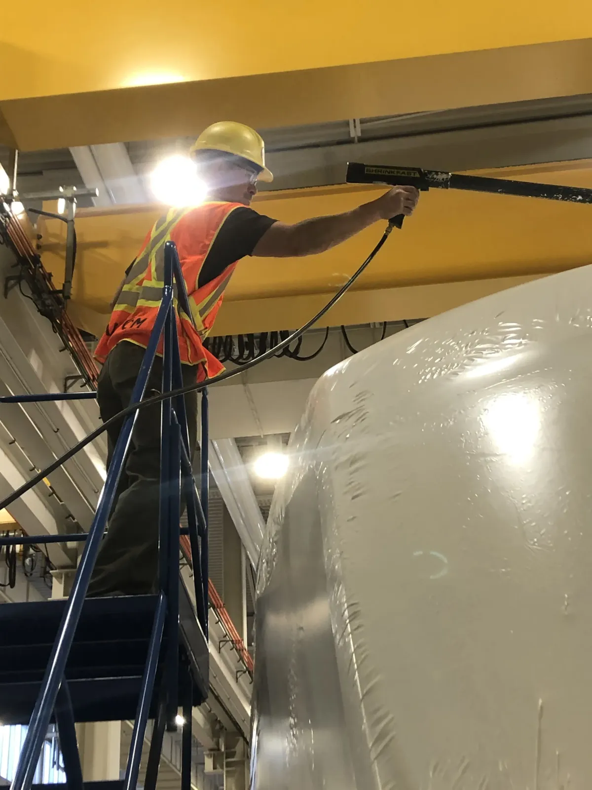 Technician on a ladder heat-sealing white shrink wrap around a large vessel inside an industrial plant