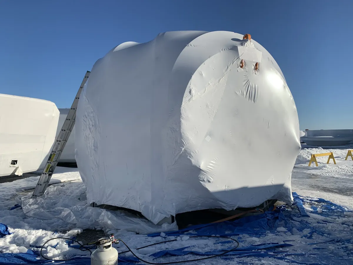 Oversized industrial vessel fully shrink wrapped in white film outdoors, too large and irregular for a shipping crate