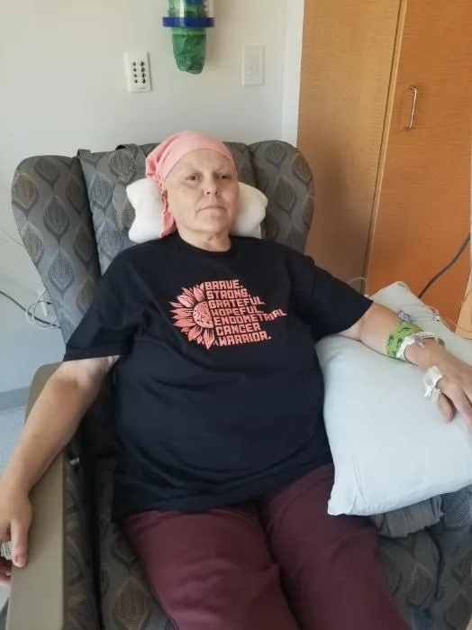 Woman undergoing cancer treatment sitting in a hospital chair wearing a pink headscarf and a black shirt that reads ‘Brave, Strong, Grateful, Hopeful — Endometrial Cancer Warrior.’