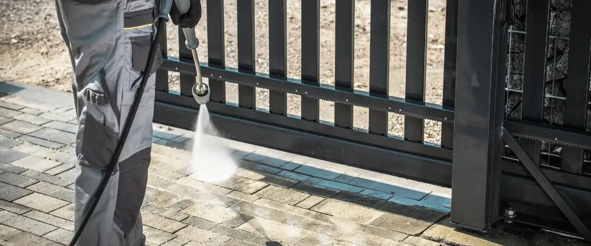 Worker using a pressure washer to clean a brick driveway near a metal gate.