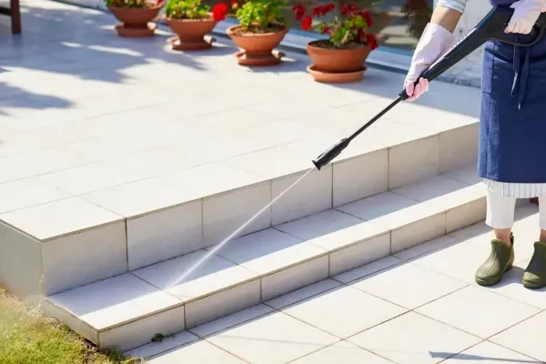An overhead view of a person in yellow wellington boots pressure washing a dirty paver pathway next to garden greenery.