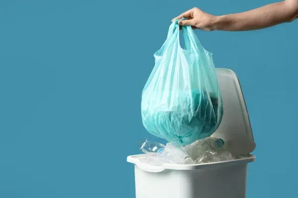 A hand holds a teal plastic bag full of recycling over a white bin with plastic bottles, against a blue background.