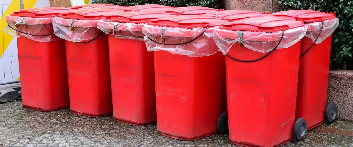 A line of six bright red garbage bins with plastic bags and locks on their lids, sitting on a cobblestone street.