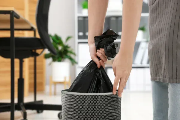 A person's hands pull a full black trash bag from a metal mesh wastebasket in a modern office.