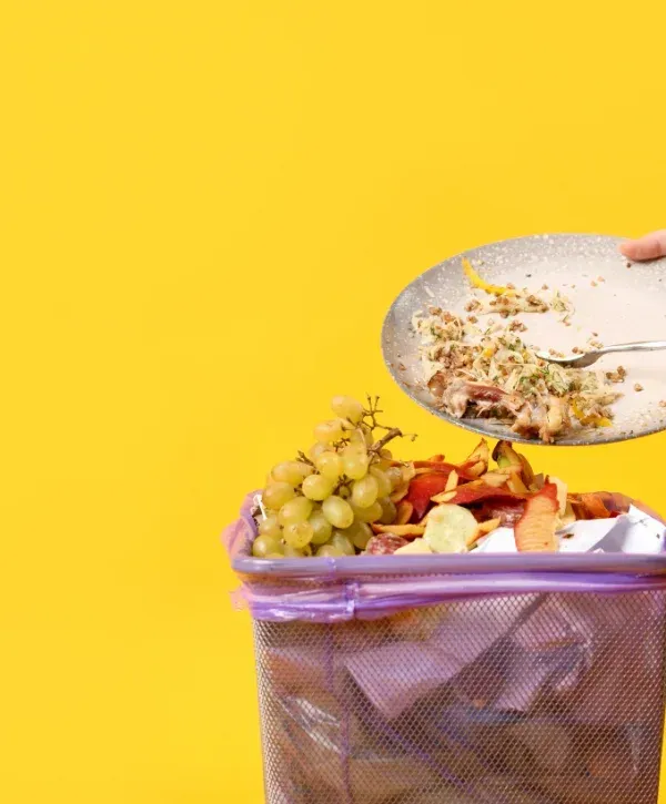 A hand scrapes leftover food from a plate into a metal trash can overflowing with food waste and a purple bag, against a yellow background.