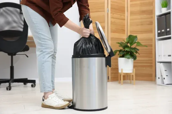 A person in light wash jeans and a brown shirt removes a full black trash bag from a stainless steel bin.