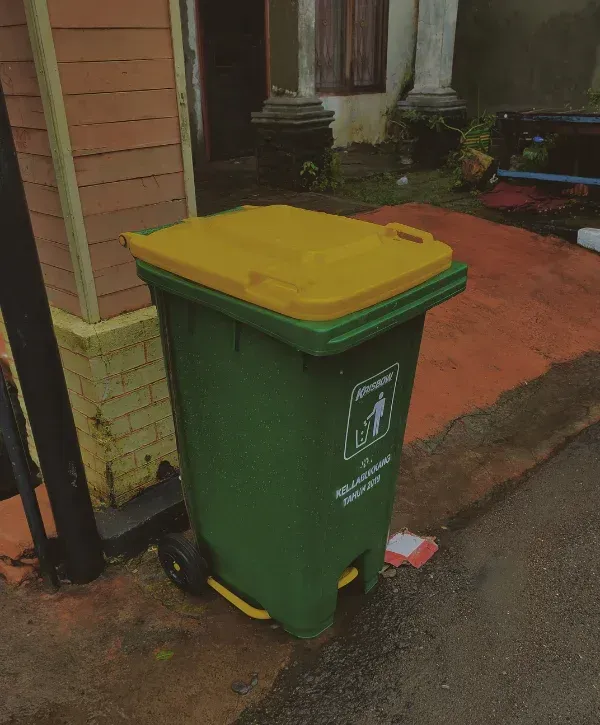 A tall green trash bin with a yellow lid and wheels stands on a wet sidewalk next to a building.