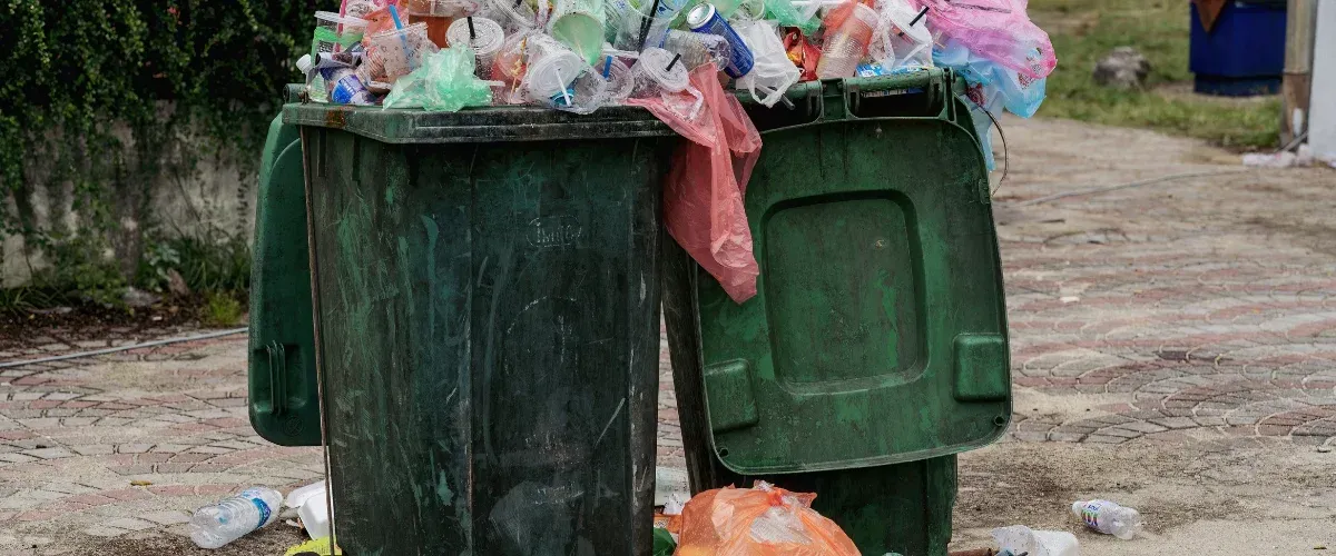 A dark green dumpster filled to overflowing with plastic waste, bags, and cups sits on a brick path.