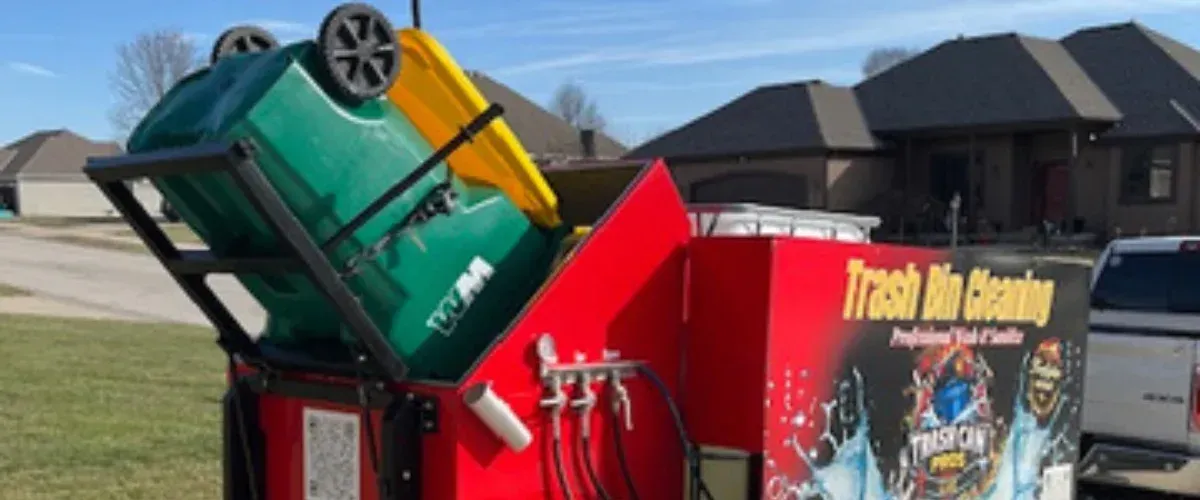 A green trash bin being lifted into a red professional trash bin cleaning machine in a residential setting.