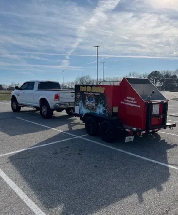 A white pickup truck towing a red trash bin cleaning trailer in a sunny parking lot under a blue sky.