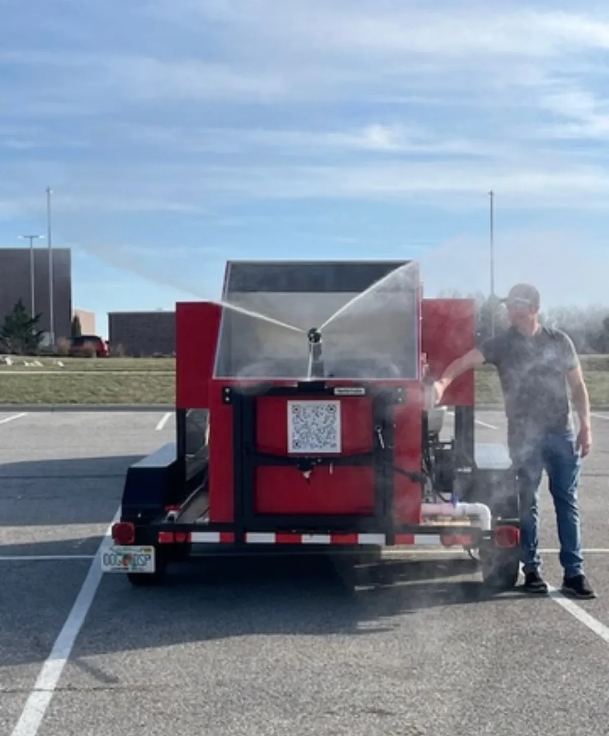 A man in a cap stands next to a red trailer-mounted mist sprayer actively spraying water in a parking lot.