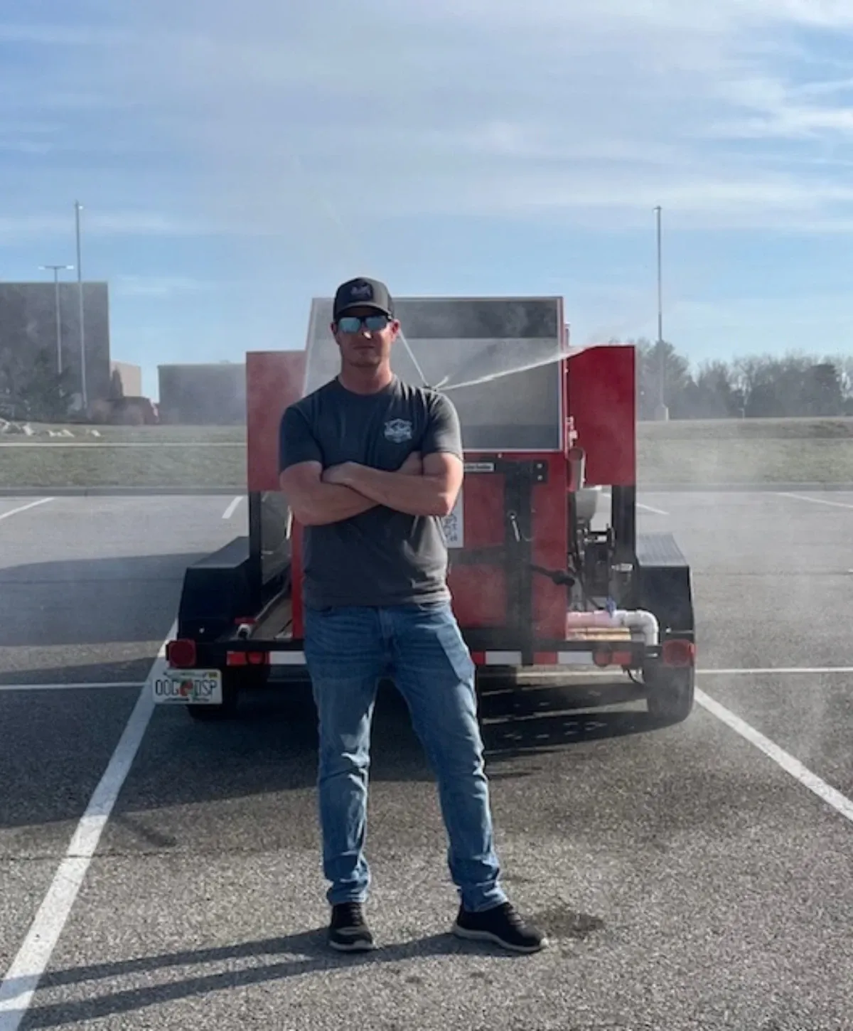 A man in a hat and sunglasses stands confidently with arms crossed in front of a red trailer-mounted spraying machine.