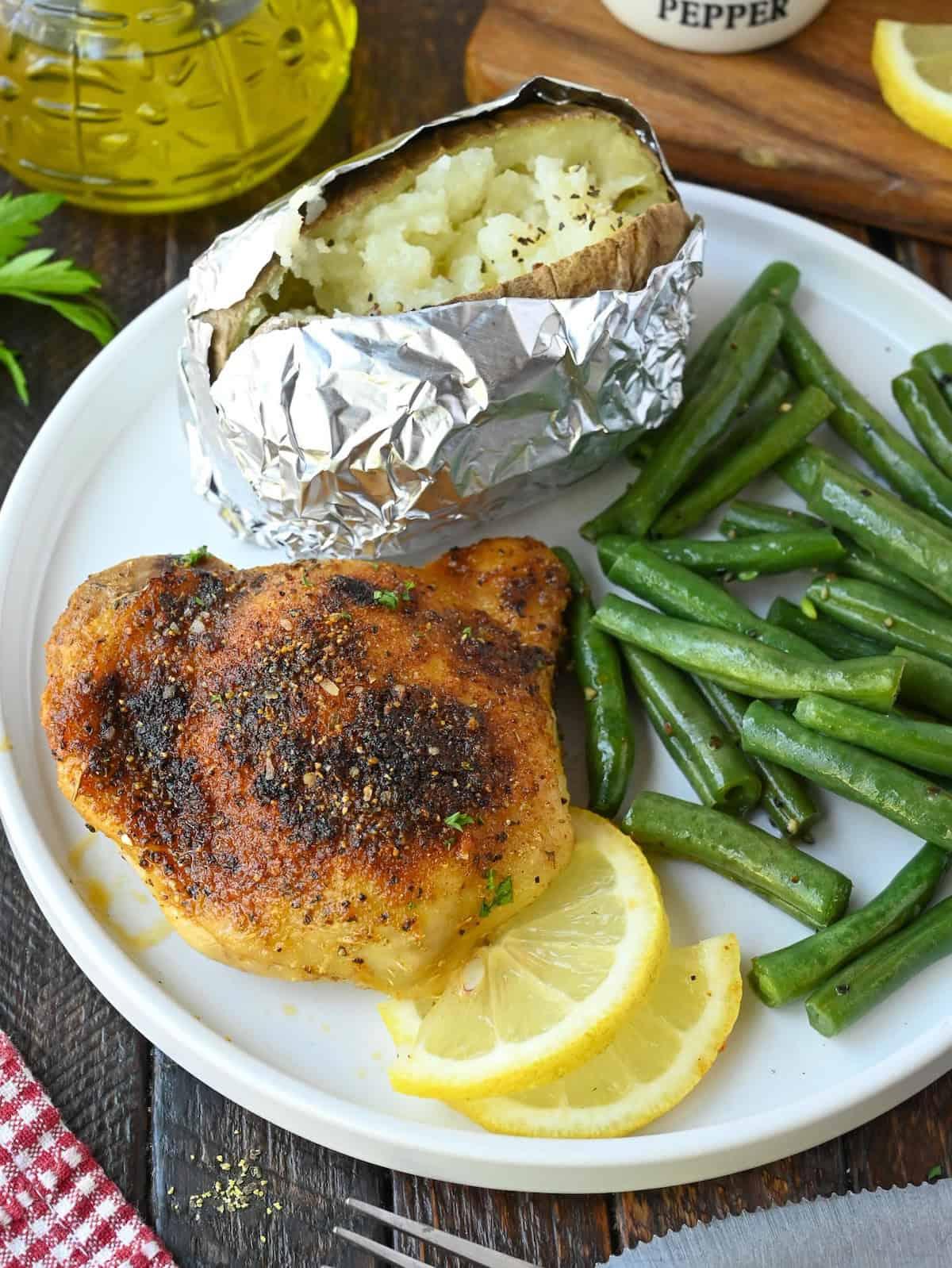 Plate with seasoned baked chicken thigh, foil-wrapped baked potato, and green beans garnished with lemon. Informative, product-related.