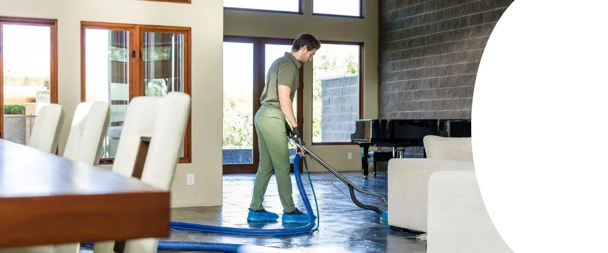 A man using a professional cleaning machine to clean the floor in a spacious living room with large windows and furniture.