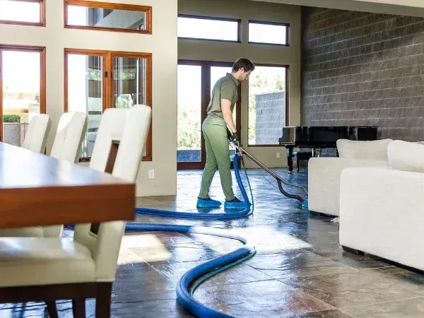 A man in a green uniform and shoe covers cleans a large stone floor in a bright, modern living room with a floor cleaner.