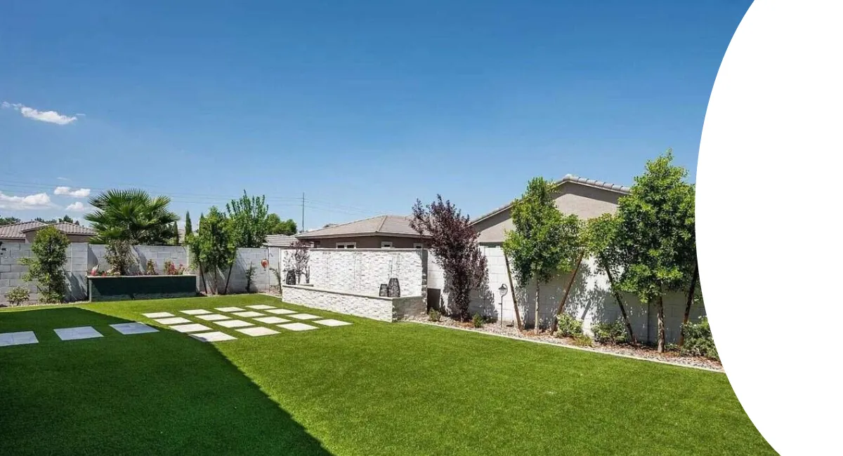 A backyard boasts a vibrant green artificial turf lawn with square stepping stone pavers and a decorative stone wall.