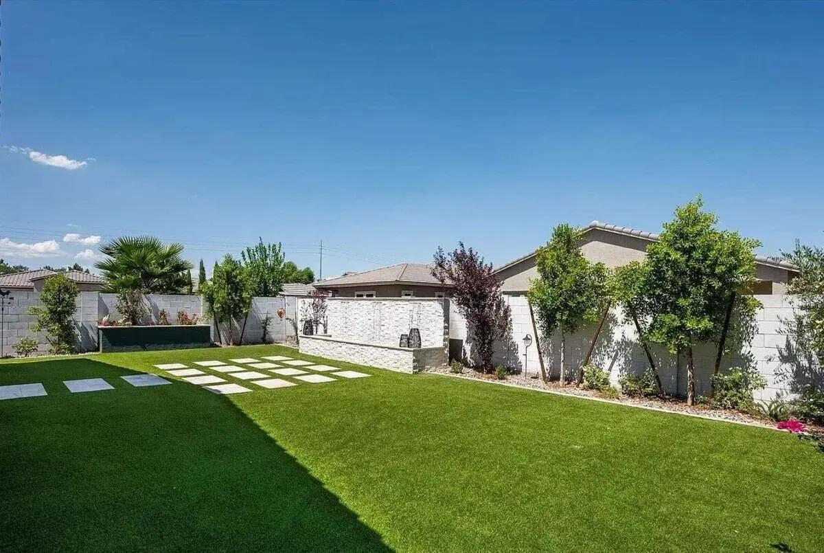 A backyard boasts a vibrant green artificial turf lawn with square stepping stone pavers and a decorative stone wall.