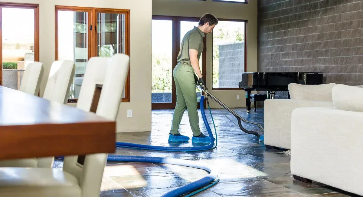 A man in a green uniform and shoe covers cleans a large stone floor in a bright, modern living room with a floor cleaner.