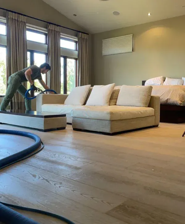 A man in green scrubs deep cleaning a light-colored sofa with a blue hose in a bedroom with wooden floors.