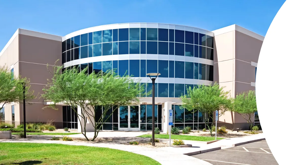 A modern office building with a curved glass facade reflecting the sky, surrounded by green landscaping and a parking lot.