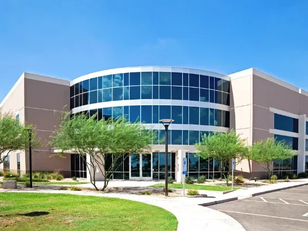 A modern curved office building with blue glass windows, beige walls, green landscaping, and a clear blue sky.