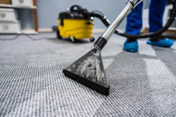 A close-up of a transparent carpet cleaning head with dirty water inside, cleaning a light gray textured carpet.