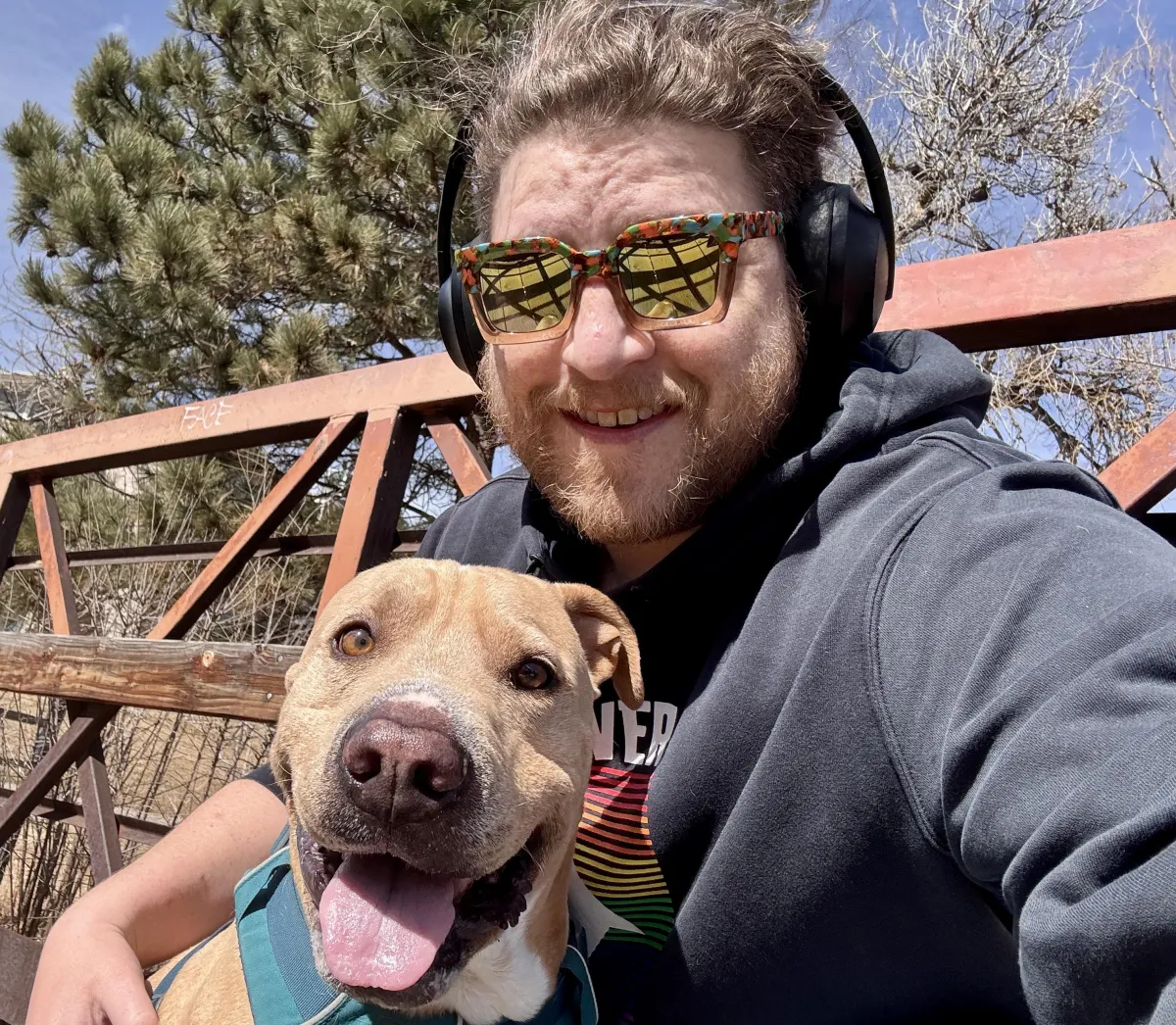 Man smiling outdoors while wearing colorful sunglasses and headphones, sitting on a rustic metal bridge with a happy tan dog in a teal harness. Bright sunny day with pine trees and blue sky in the background.