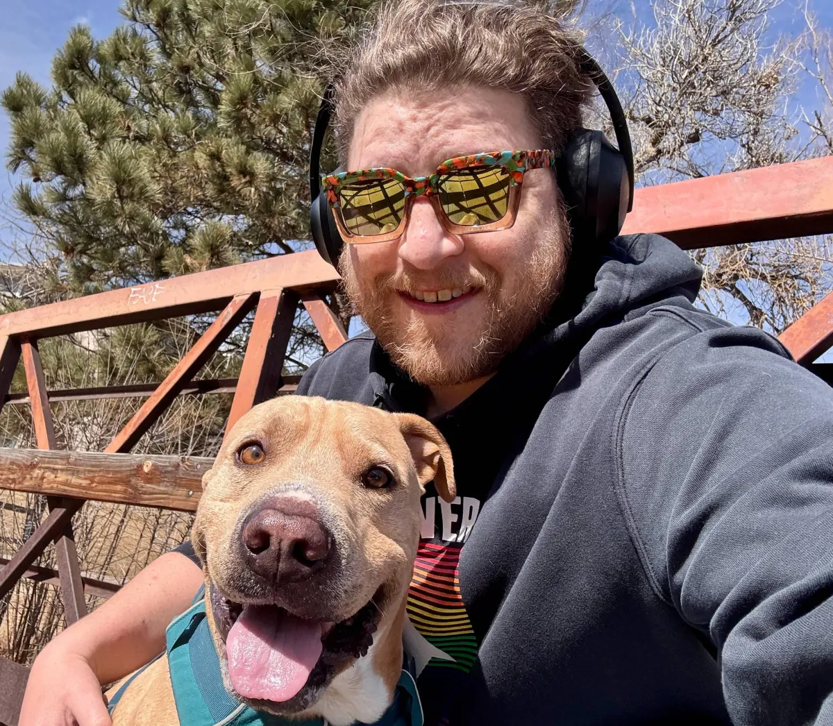 Solenzo owner smiling outdoors while wearing colorful sunglasses and headphones, sitting on a rustic metal bridge with a happy tan dog in a teal harness. Bright sunny day with pine trees and blue sky in the background.