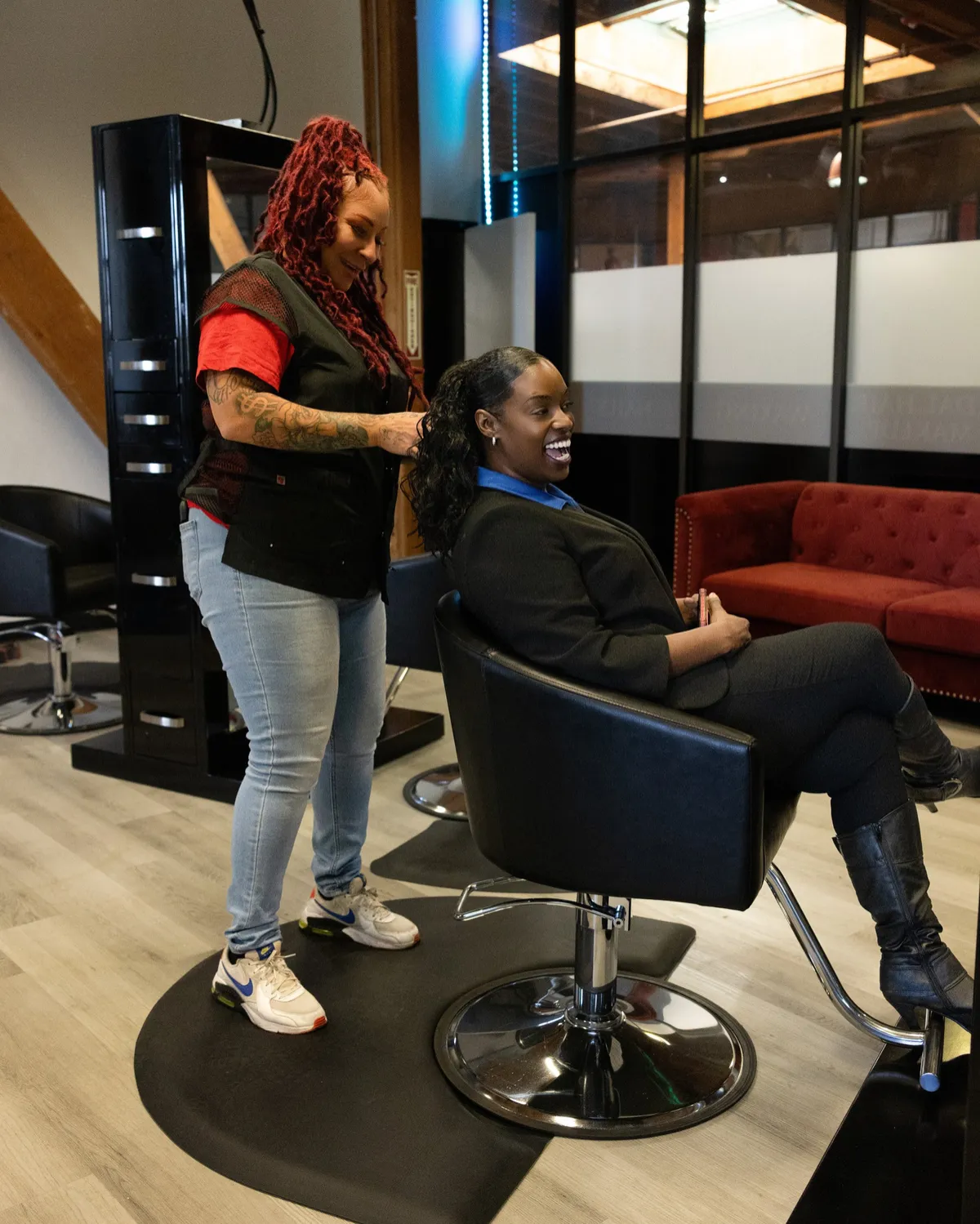 A client laughs while her Style City stylist works on her hair, red velvet sofa behind