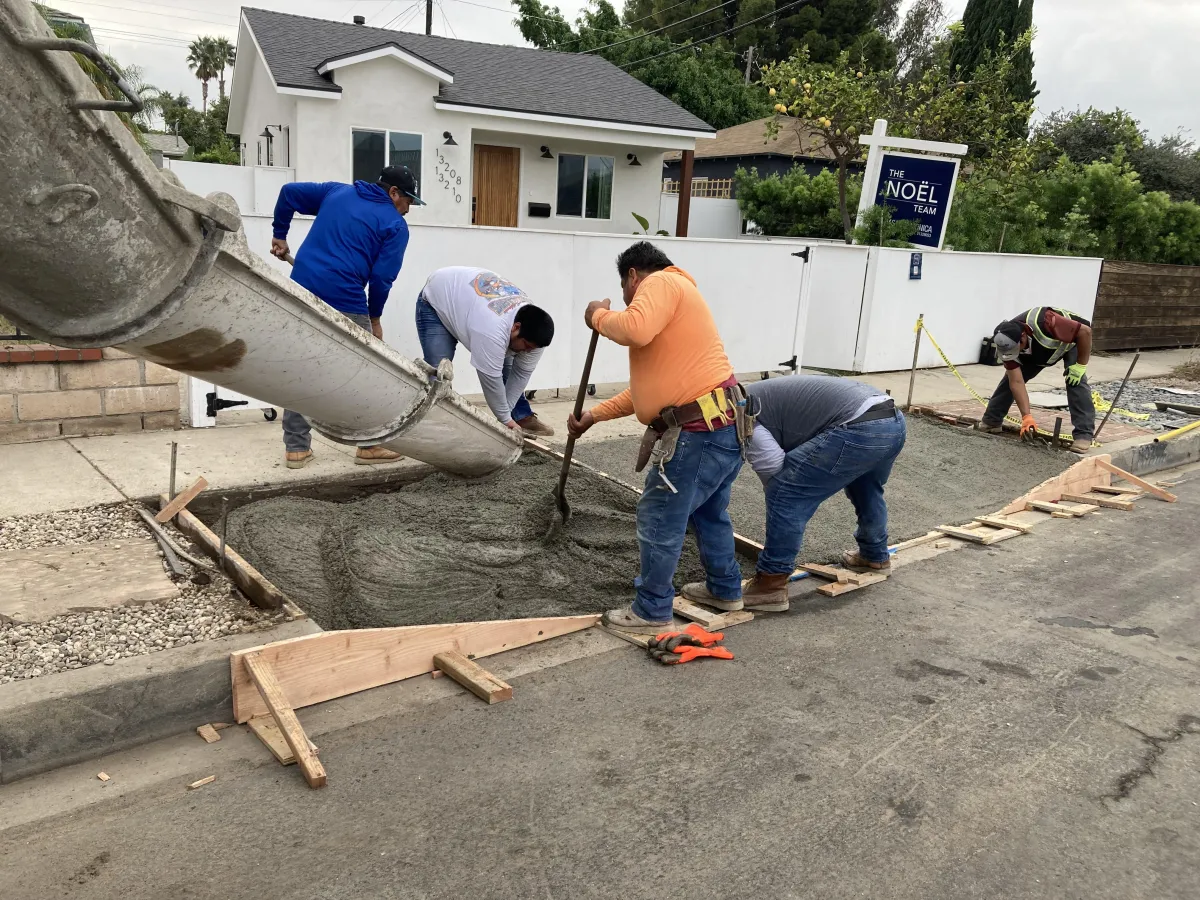 Image shows a crew of 5 workers pouring concrete directly out of dhe mixer truck into a formed driveway approach .