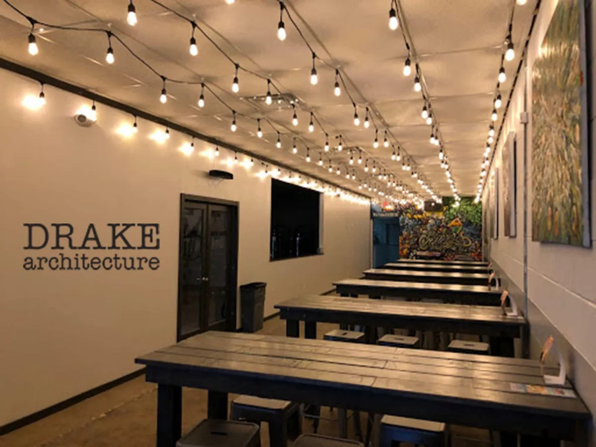 Interior view of Alematic Brewery’s seating area featuring industrial-style lighting, communal wooden tables, and vibrant street art on the walls, showcasing the design work by Drake Architecture.