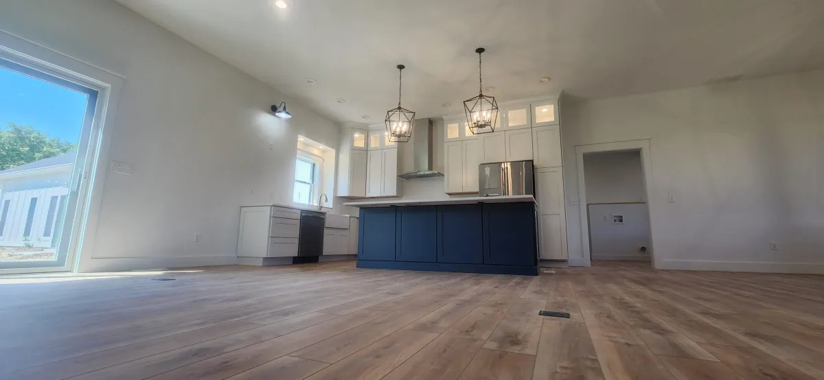 Spacious modern kitchen with an open layout, featuring light wood floors, navy blue island, white cabinetry, and large windows providing natural light.