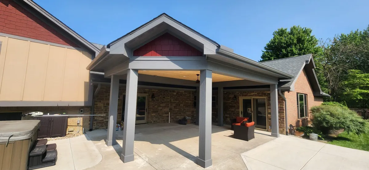 Patio addition to a brick house, featuring a covered roof supported by columns, with stone accents and comfortable seating, designed to enhance outdoor living space.