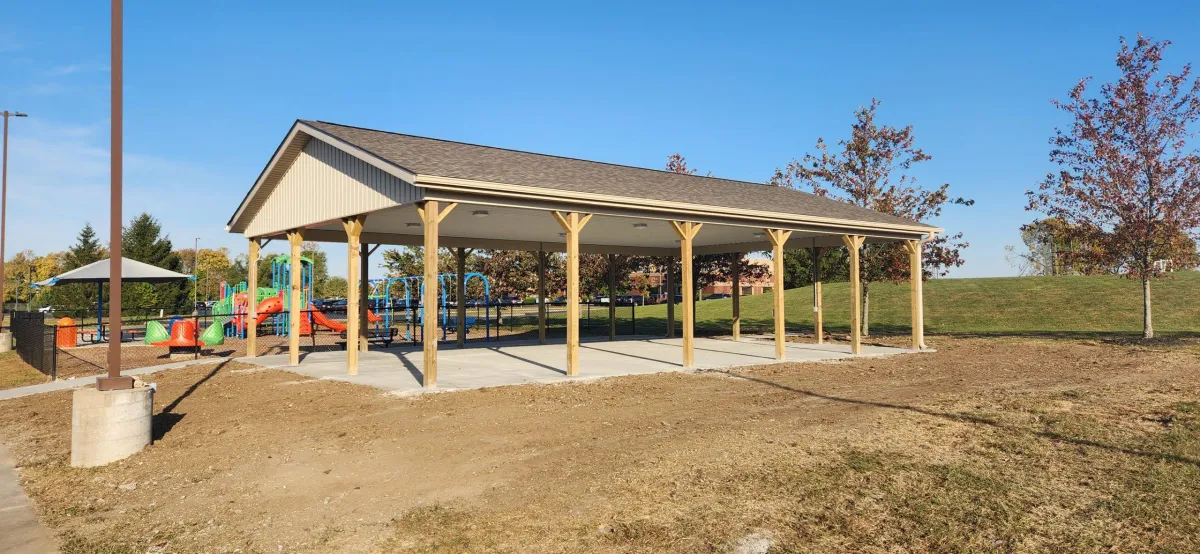 Large covered pavilion in a community park, situated next to a playground, featuring a pitched roof and open sides supported by wooden columns.