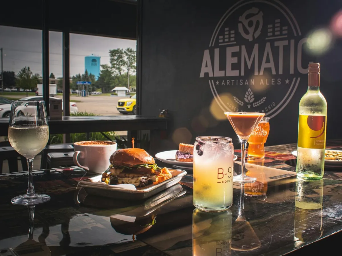 Close-up of a dining setup at Alematic microbrewery in North Dayton, featuring a burger, cocktails, and wine on a polished table with the Alematic logo in the background, showcasing the industrial aesthetic design by Drake Architecture.