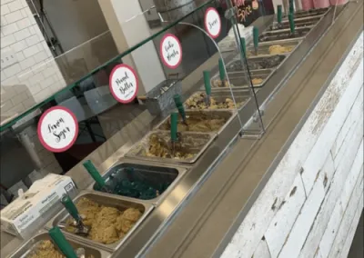 Close-up of the cookie dough display at No-Baked Cookie Dough shop, featuring various flavors in metal trays behind a glass barrier, with rustic whitewashed wood counter, part of the modern design by Drake Architecture.